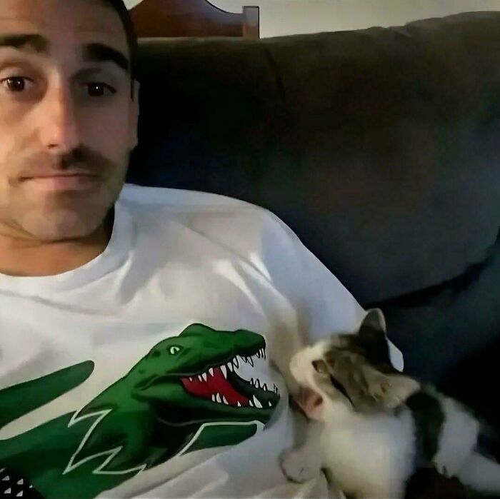 A peculiar photo of a man on a couch, looking at the camera, while a small kitten naps on his chest.