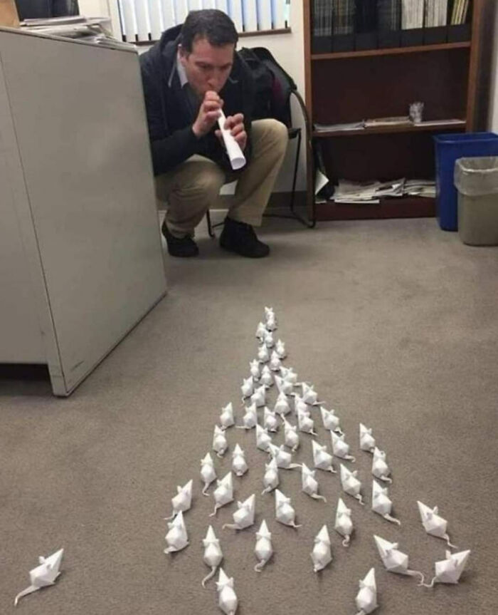 A man crouching, blowing through a paper tube towards a formation of peculiar origami mice on the floor. Spotted in real life.