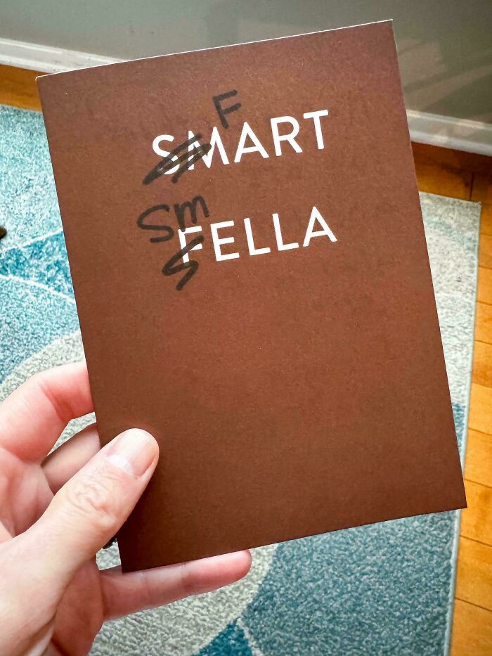 A hand holds a brown card with SMART FELLA, showing crossed out letters. Dads will find this relatable.