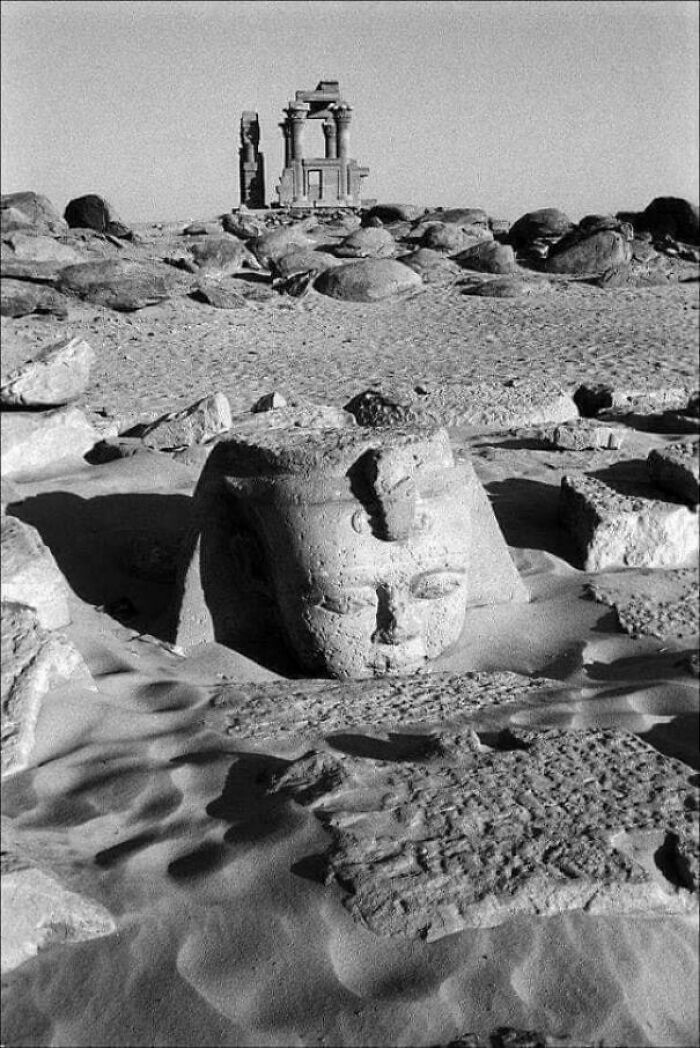 The ancient head of a sphinx partially buried in desert sand with ruins in the background, a very interesting photo.