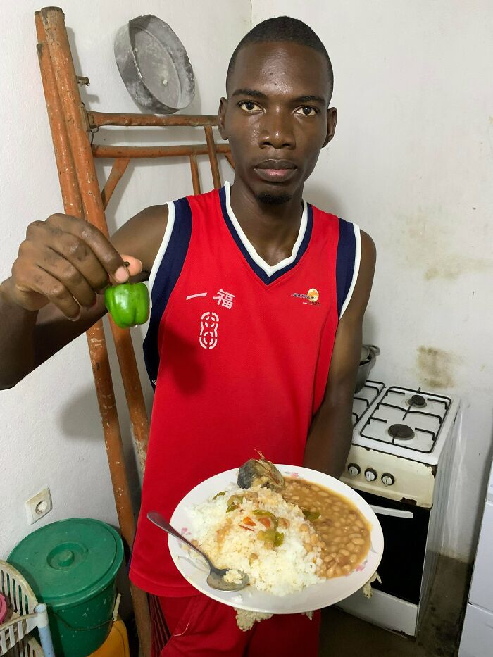 A man holding a tiny green pepper and a plate of food (rice, beans, fish) in a kitchen, from the Not Interesting community.