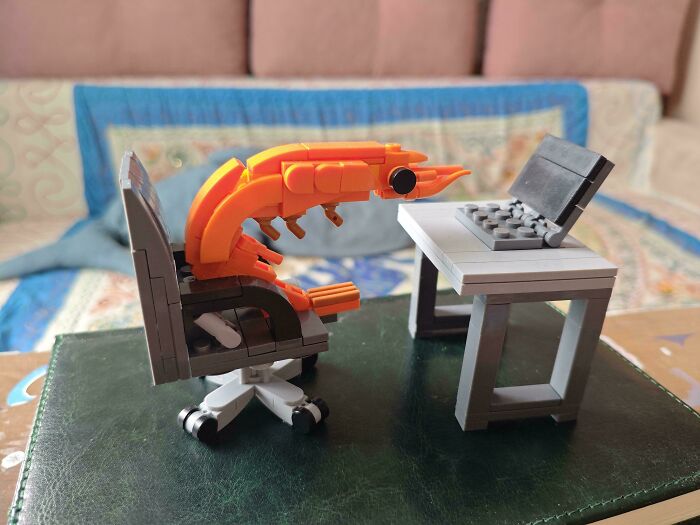 A LEGO shrimp character sitting on a LEGO office chair in front of a LEGO desk with a laptop, from the Not Interesting community.