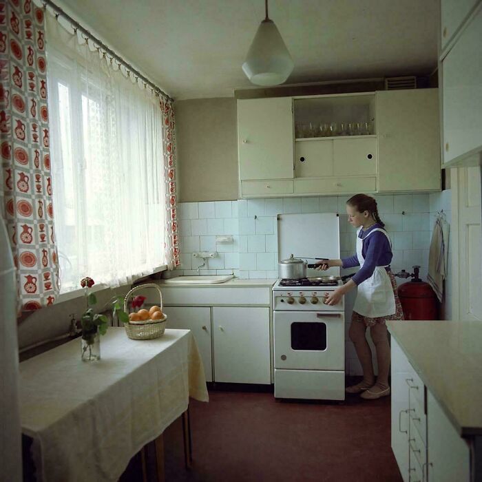 A historical photo of a young girl cooking in a retro-style kitchen with patterned curtains and white cabinets, showing typical days we will never experience.