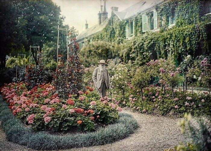 A colorized historical photo of a man in a hat standing in a vibrant flower garden, with a large house in the background, a scene from days we will never experience.