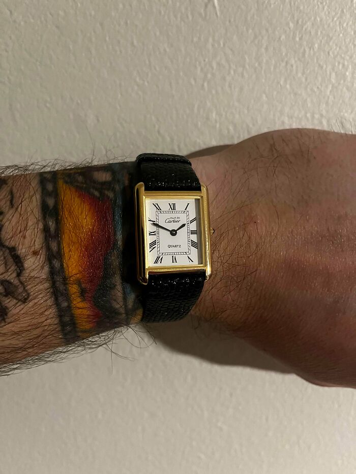 A gold Cartier watch with a black leather band, a thrift store jackpot, on a person's tattooed wrist.