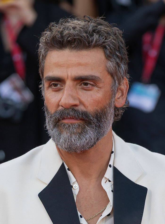 Oscar Isaac with salt and pepper beard wearing a white jacket and polka dot shirt at a formal event.