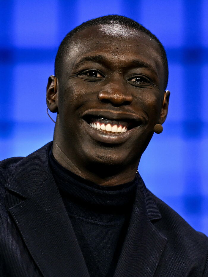 Khaby Lame smiling during a speaking event, wearing a black turtleneck and jacket with a microphone headset.