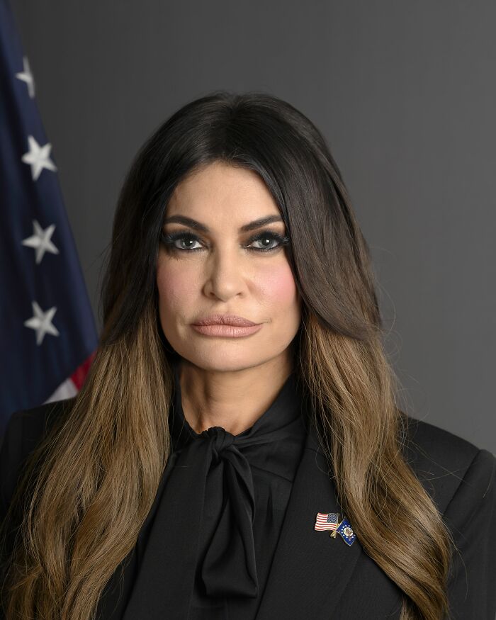 Kimberly Guilfoyle with long hair and black outfit, posing formally with US flag in the background for career highlights.