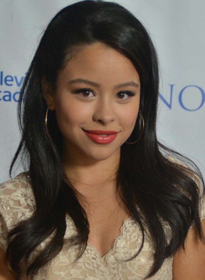 Cierra Ramirez smiling at an event, wearing gold hoop earrings and a lace beige top with long dark hair.