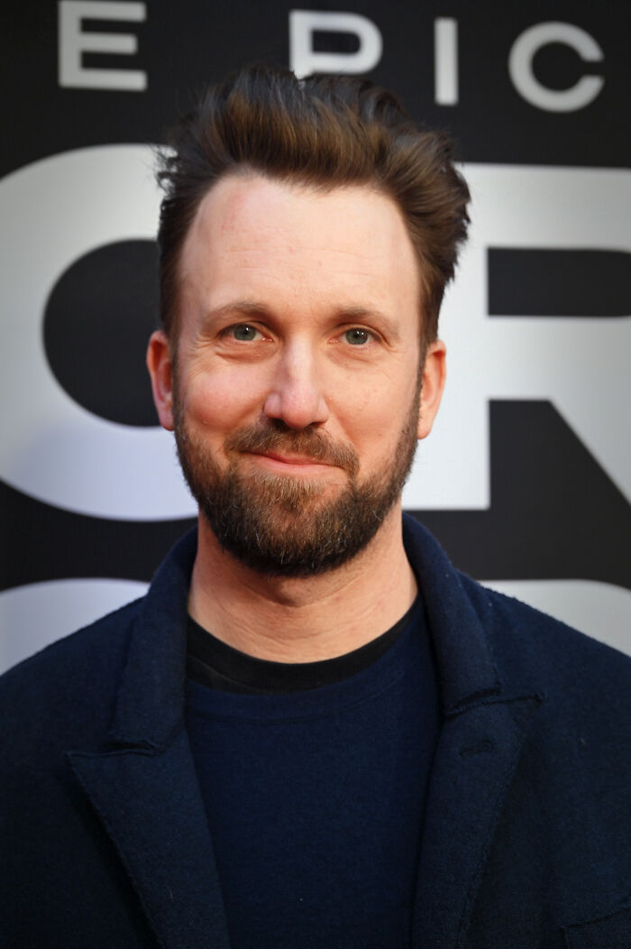 Jordan Klepper with beard and dark jacket smiling at event, posing against black and white backdrop.