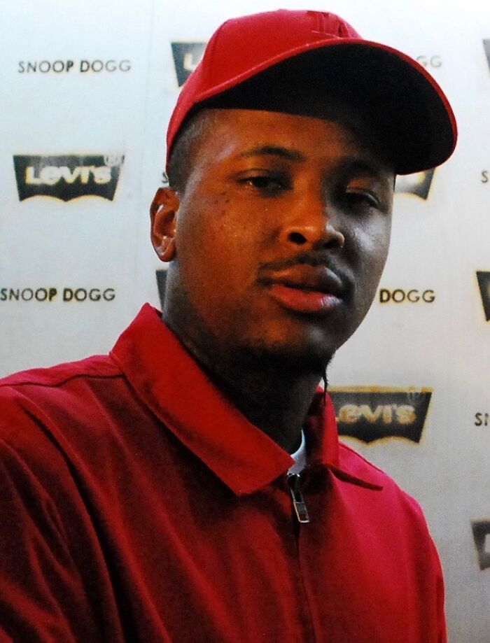 YG in a red cap and jacket at a promotional event with Levi's and Snoop Dogg logos in the background, showcasing career highlights.