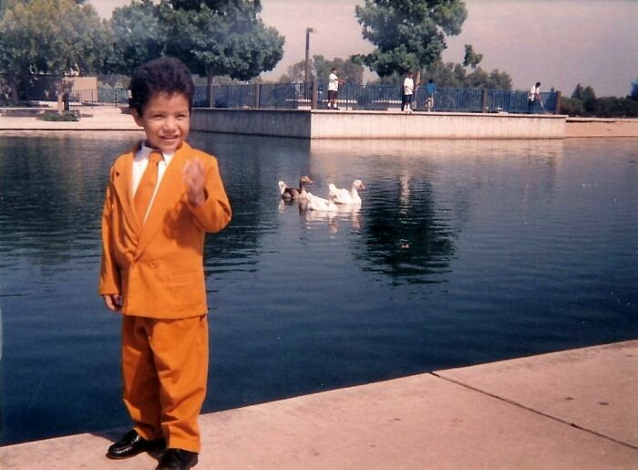 Young boy in an oversized orange suit standing by a pond with ducks, a hilariously awkward childhood pic.