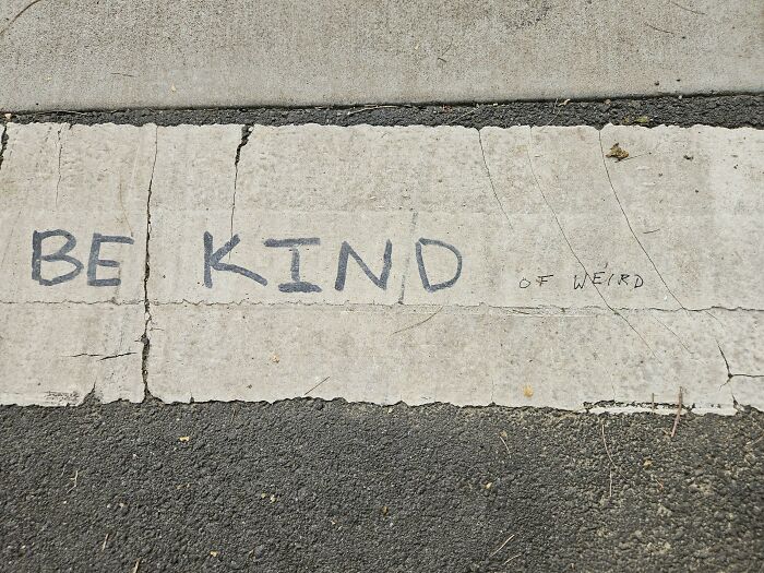 Mild vandalism graffiti on a cracked concrete surface with the words Be Kind of Weird written in black marker.
