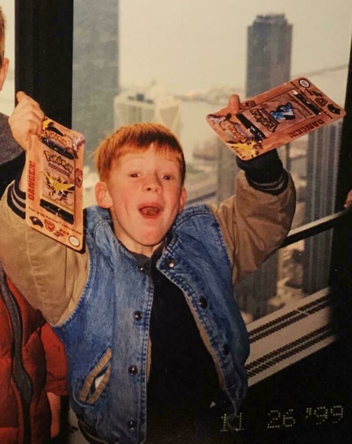 Childhood photo shows excited kid holding vintage Pokémon cards in a hilariously awkward pose indoors by a city window.