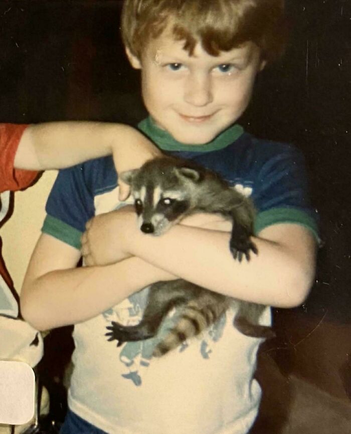 Young boy holding a raccoon with a mischievous smile, a classic awkward childhood moment captured in a vintage photo.