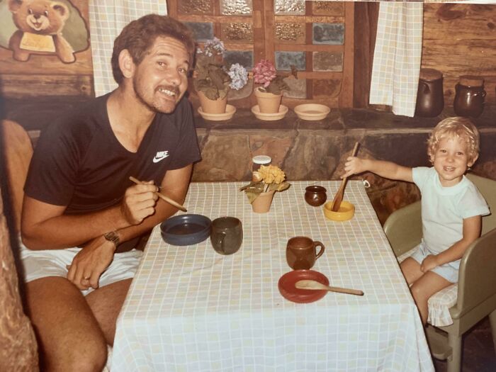Man and child posing with wooden spoons at table in a rustic kitchen, a hilariously awkward childhood pic shared online.
