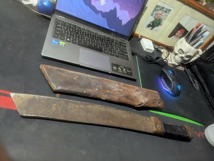 Old rusty machete with worn leather sheath found in an old home beside a laptop and skull-shaped pen holder.
