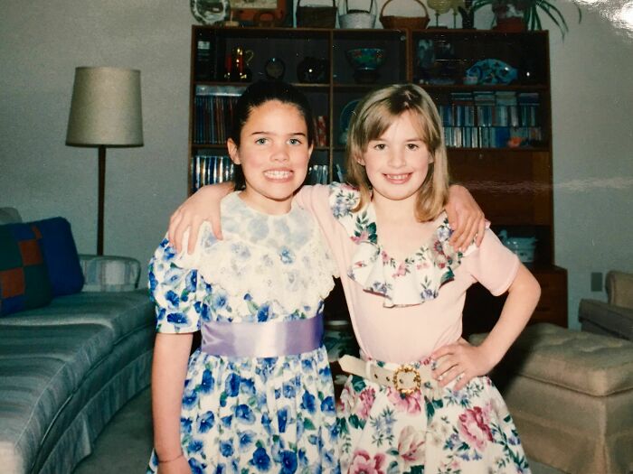 Two young girls in floral dresses smiling awkwardly, capturing a hilariously awkward childhood moment to share and remember.