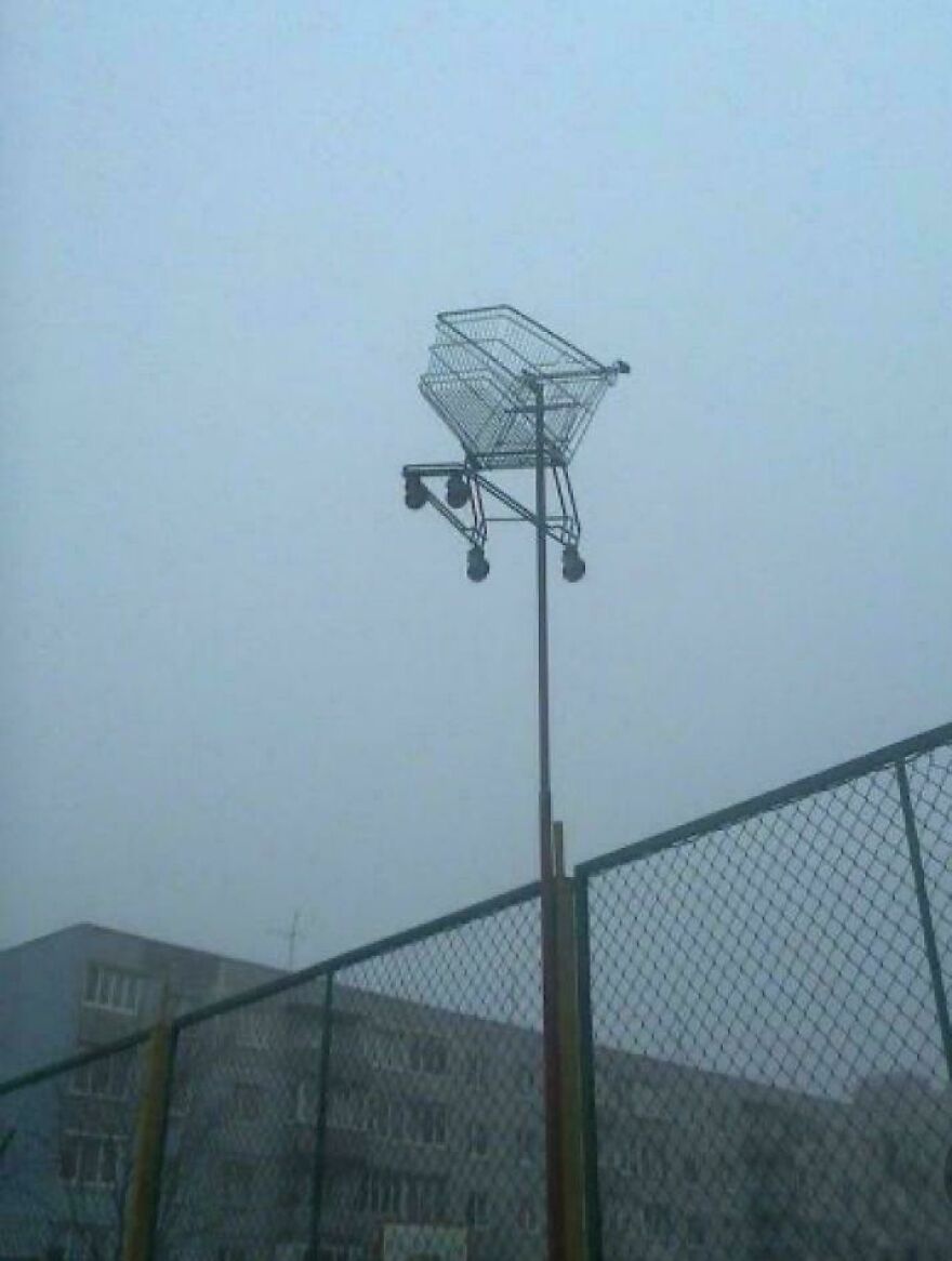 Weird and unexplainable sight of a shopping cart inexplicably stuck high atop a pole near a fenced area.