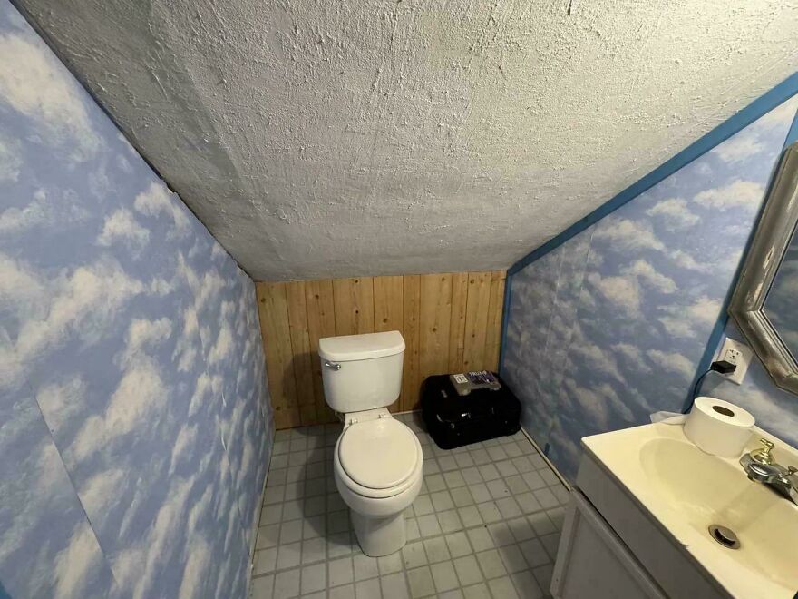 Small bathroom with low slanted ceiling, sky wallpaper, and a toilet placed unusually close to the wood-paneled wall.