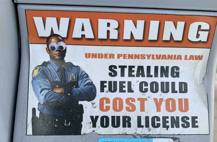 Warning sign about stealing fuel altered with googly eyes on police officer, a mild vandalism example to cheer you up.