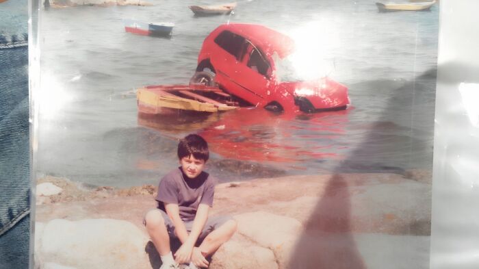 Childhood photo of a young boy sitting near water with a red car half submerged in the background, awkward and funny moment.