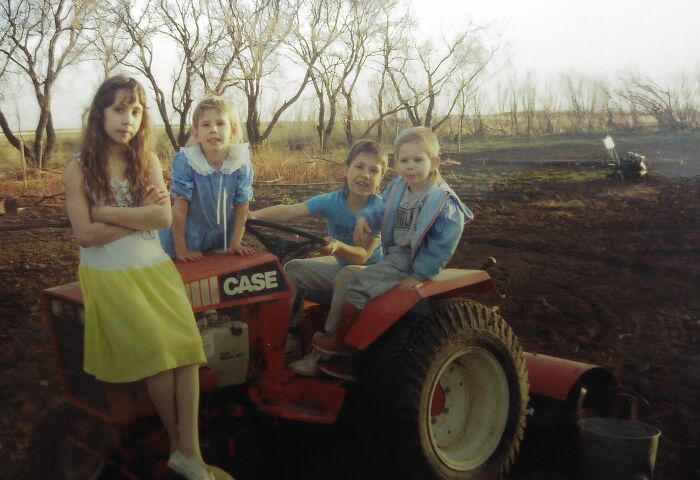 Four children posing awkwardly on and around an old tractor in a barren field, captured in a hilariously awkward childhood pic.