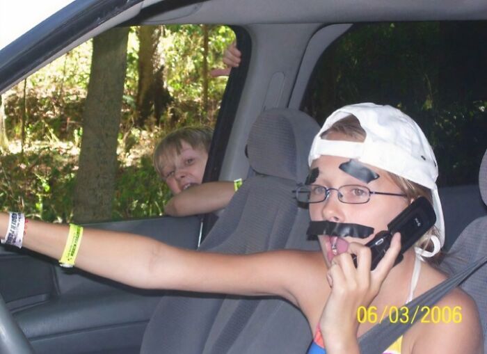 Child wearing fake mustache and glasses on phone in car while another child leans out window in awkward childhood pics.