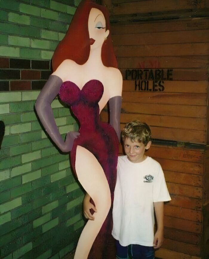 Young boy posing awkwardly next to a large Jessica Rabbit statue, a hilariously awkward childhood moment.