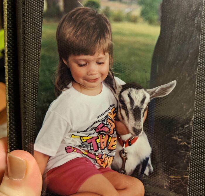 Child with a mullet hairstyle wearing a graphic tee sitting outdoors holding a small goat, a hilariously awkward childhood pic.