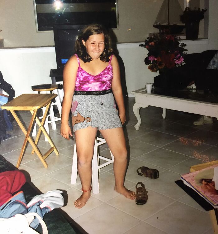 Girl in a pink top and shorts making a funny face while sitting awkwardly indoors in a hilariously awkward childhood pic.