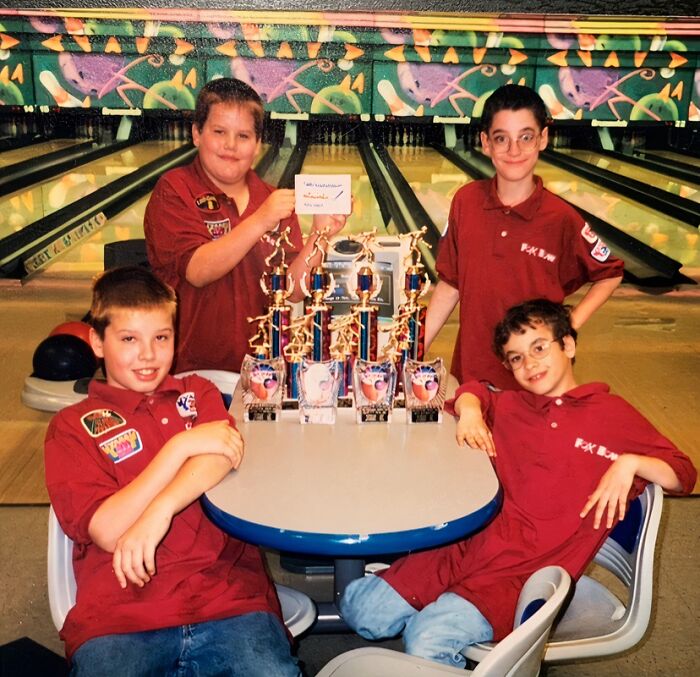 Four kids in matching shirts at a bowling alley posing with multiple trophies in a hilariously awkward childhood photo.
