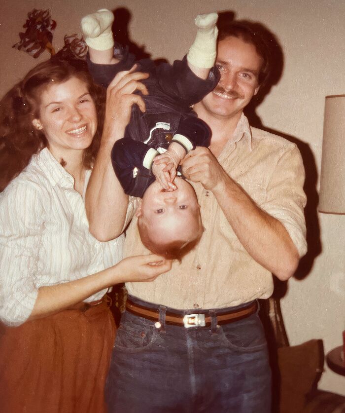 Smiling parents holding an upside-down baby in a vintage room, capturing a hilariously awkward childhood moment.