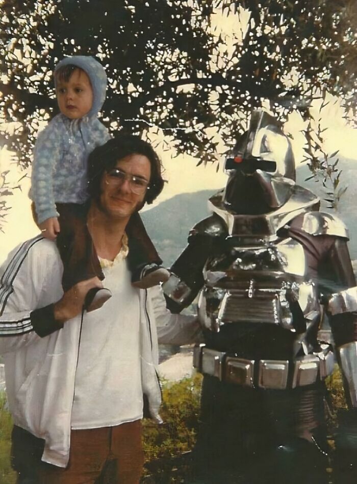 Man carrying a child on his shoulders posing with a person in a shiny robot costume in an outdoor setting, awkward childhood pic.