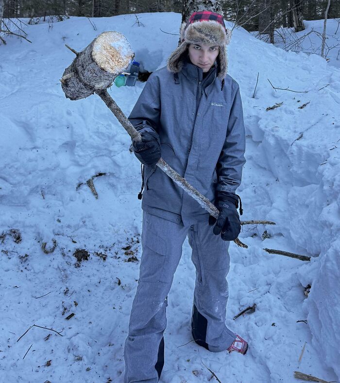 Person in winter gear holding a large cool stick made from a tree branch in a snowy forest setting.