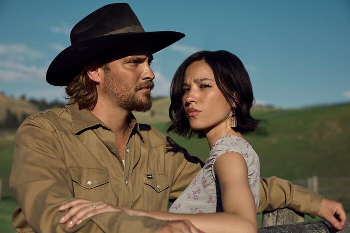 Two Yellowstone actors in western attire stand outdoors with scenic hills in the background, hinting at an infamous location.