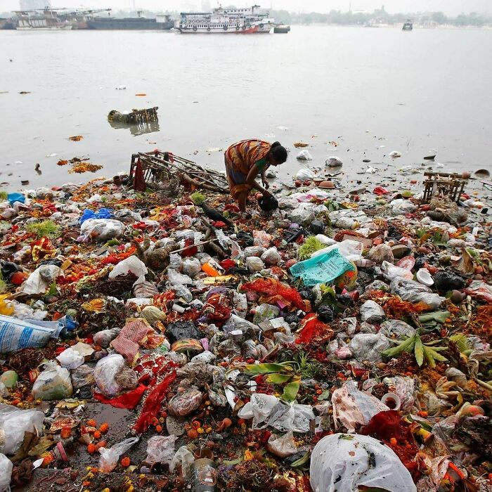 Urban hell photo of woman searching through a large pile of trash dumped by a river with boats in background