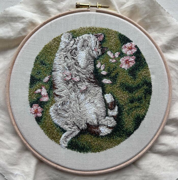 Stunning embroidery creation of a white cat sleeping on grass with pink flowers, inspiring embroidery hobby.