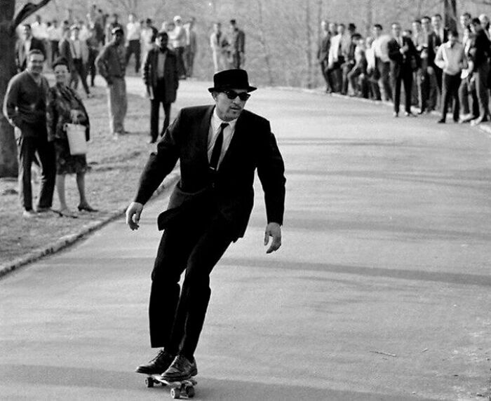 Man in a suit and hat skateboarding on a road with a crowd watching, showcasing strange old photographs concept.