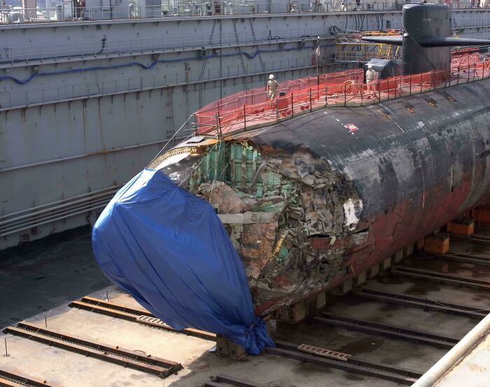 Submarine with extensive damage to its bow, covered by a blue tarp, in a dry dock. A serious accident.