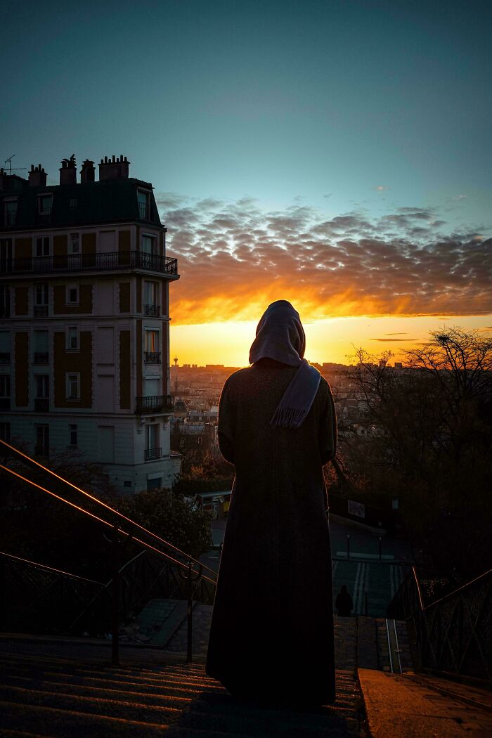 A cloaked figure standing on steps looking at a dramatic sunset over a city, showing Accidental Renaissance vibes