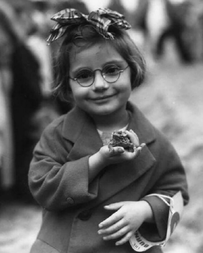 Candid old photos: A young girl wearing glasses and a bow in her hair, holding a frog in her hand.