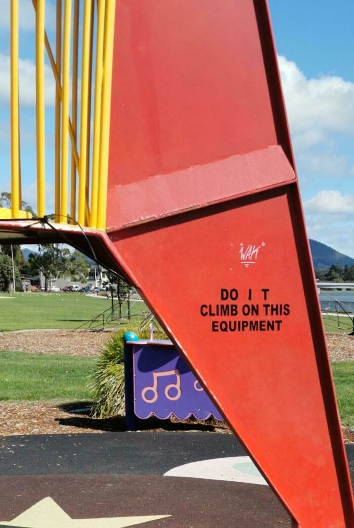 Mild vandalism on playground equipment with altered warning sign encouraging climbing in a park setting.