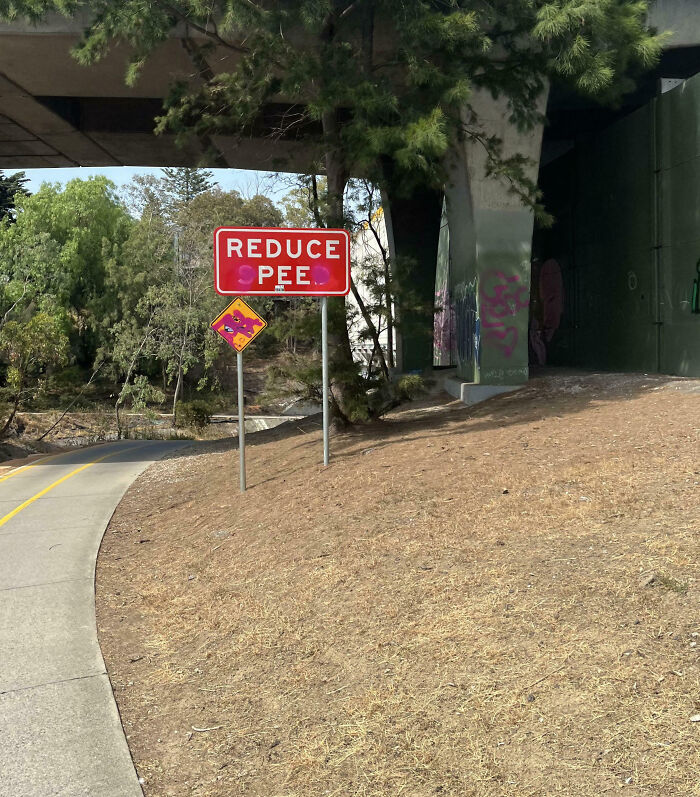 Road sign altered by mild vandalism to read reduce pee, with graffiti on nearby pillars under a bridge.