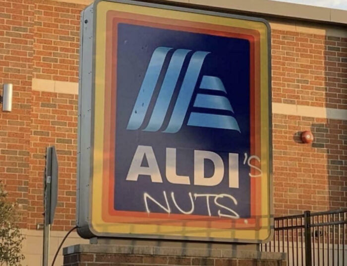 Street sign with mild vandalism on an Aldi logo, featuring graffiti that reads nuts on the brick building background.