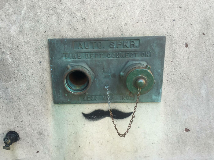 Mild vandalism showing a fire department connection with added graffiti mustache under the metal fixture on a concrete wall.