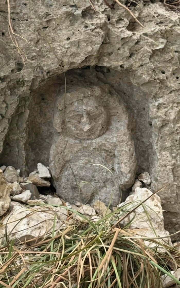Stone carving of a small creature resembling a bear or owl, partially hidden in rocky terrain with grass nearby.