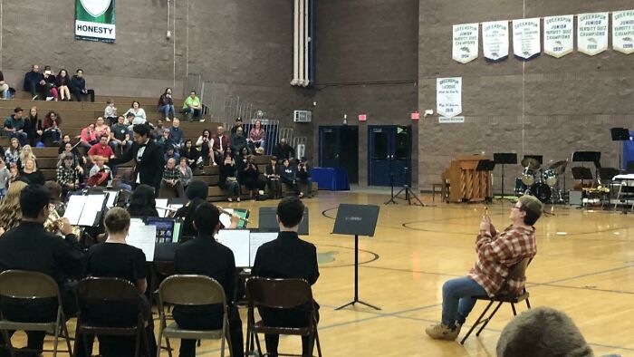 Audience watching a school band performance in a gymnasium with banners and musical instruments in the background.