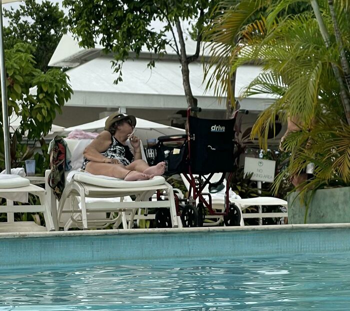 Woman relaxing on a lounge chair by the pool surrounded by tropical plants and a wheelchair, illustrating entitled Karens behavior.