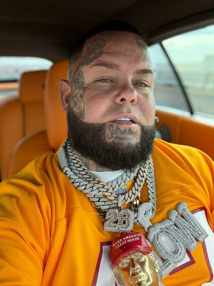 A man with a heavily tattooed face and neck, wearing multiple large chains, displaying an unfortunate haircut.